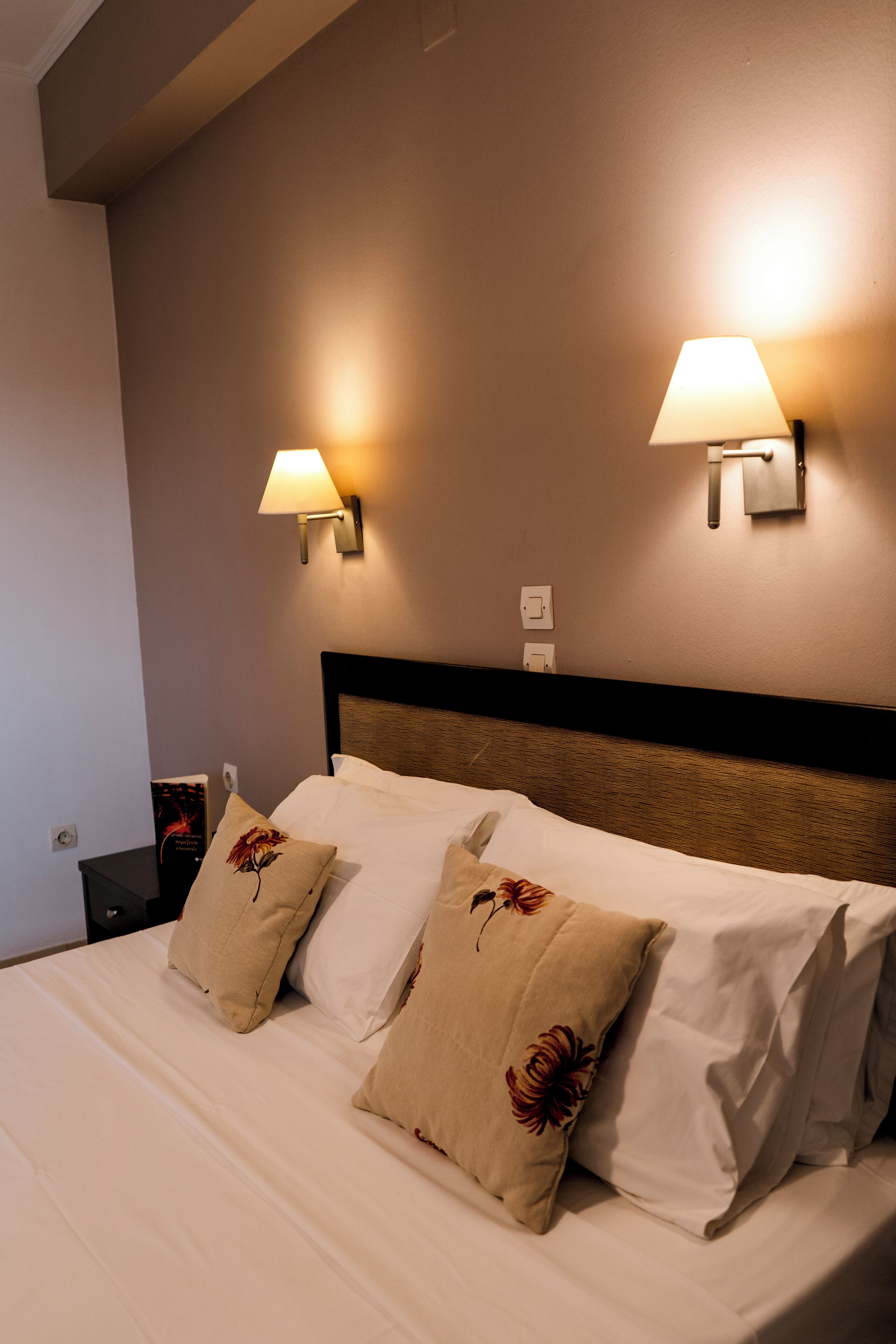 Una habitación de hotel con una cama bien hecha, dos lámparas fijadas a la pared y una mesita de noche con un libro.