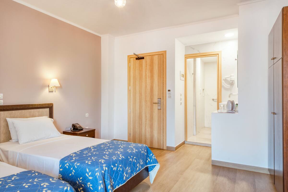A hotel room with two beds, a wooden wardrobe, a bathroom with a mirror, and a small table with a kettle.