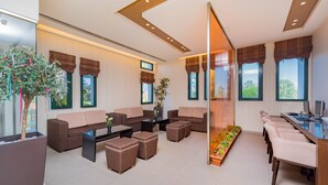 A modern living room with sofas, a coffee table, and a long table with chairs. There are large windows with curtains, a plant, and a view of the outdoors. - Oscar Hotel (Zakynthos)