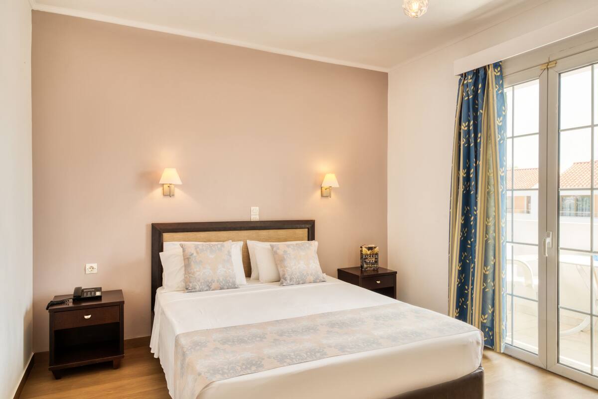 A hotel room with a bed, two bedside lamps, a nightstand, and a view through a glass door.