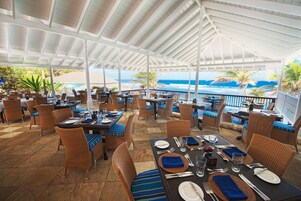 Un espacio de comedor al aire libre con mobiliario de mimbre, mesas dispuestas para dos personas y vistas al océano.