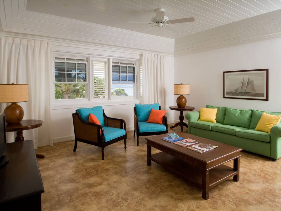 A living room with two armchairs, a green sofa, a coffee table, and a television on a stand. There are two windows with curtains, a ceiling fan, and a framed picture on the wall.