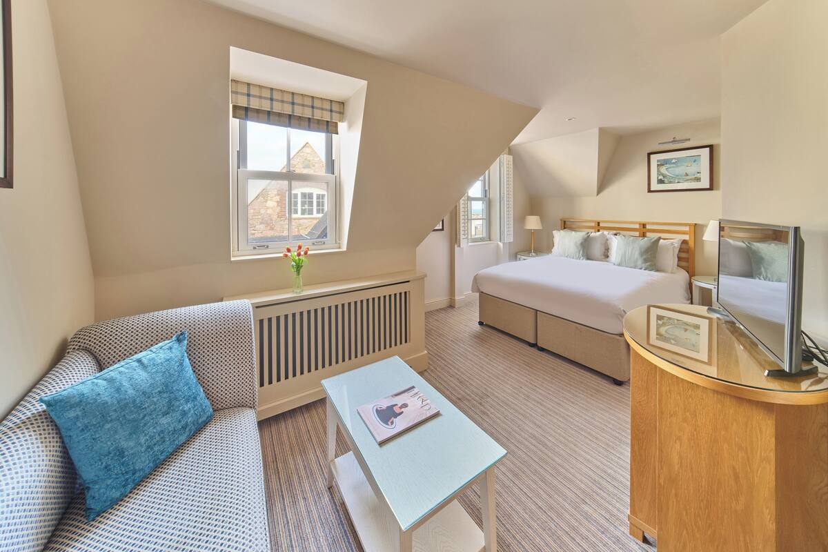 A hotel room with a bed, a sofa, a small table, a TV, and a window with a view of a building.