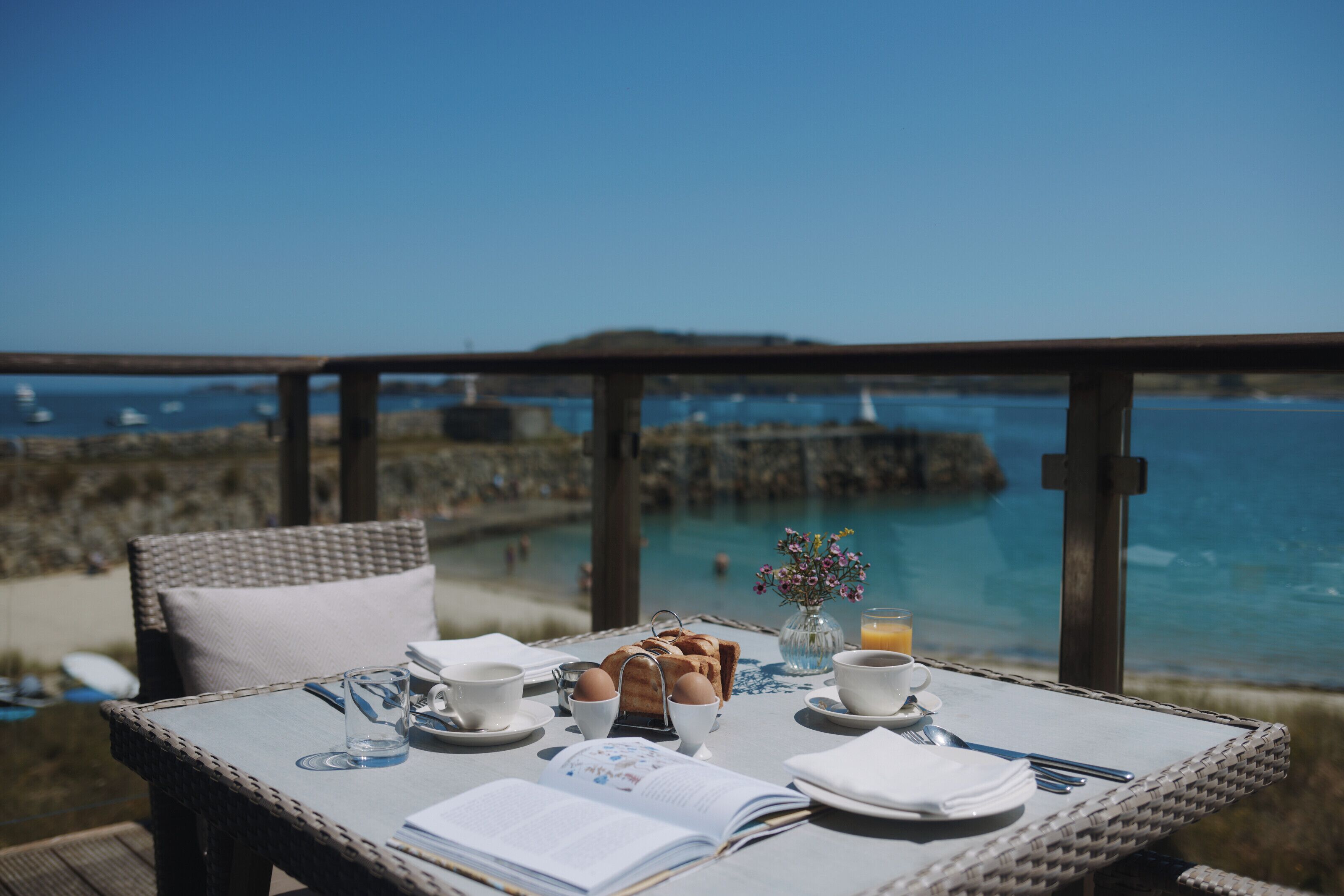 Ein Esstisch mit Frühstücksangebot, einem Buch und Blick auf das Meer.