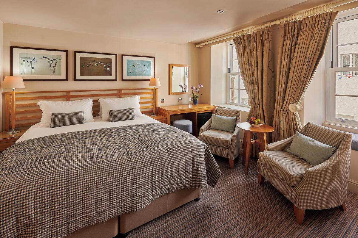 A hotel room with a large bed, two armchairs, a desk with a computer, and a window with curtains.