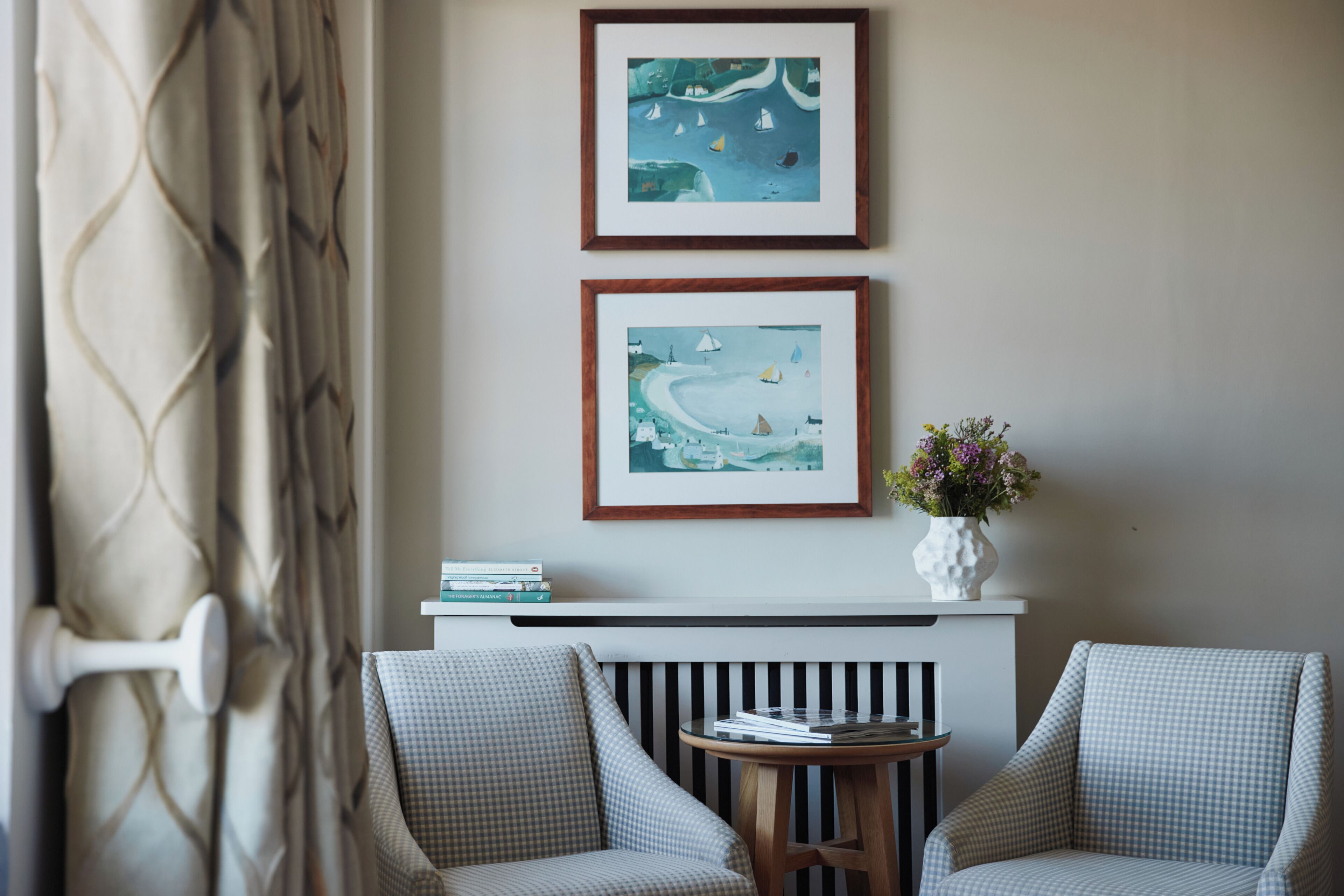 A room with two checkered armchairs, a small table with books, a white vase with flowers, and two framed artworks above a white console table.