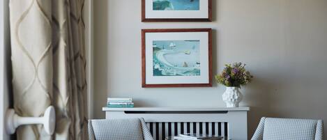 A room with two checkered armchairs, a small table with books, a white vase with flowers, and two framed artworks above a white console table.