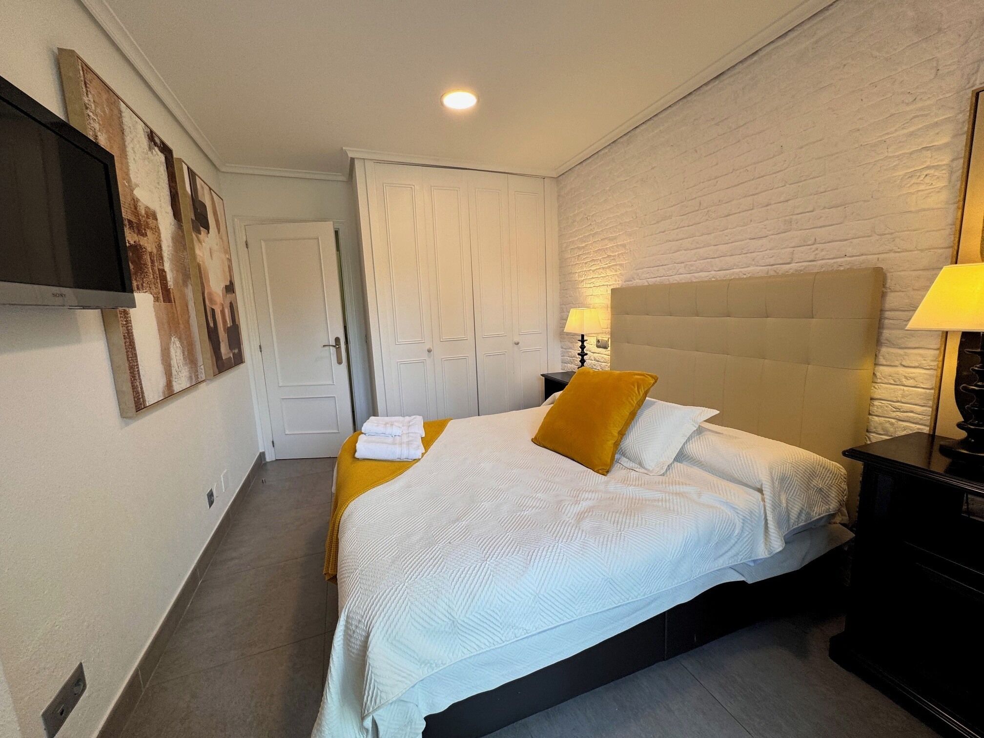A bedroom with a bed, a nightstand, a lamp, a wall-mounted TV, and a door.