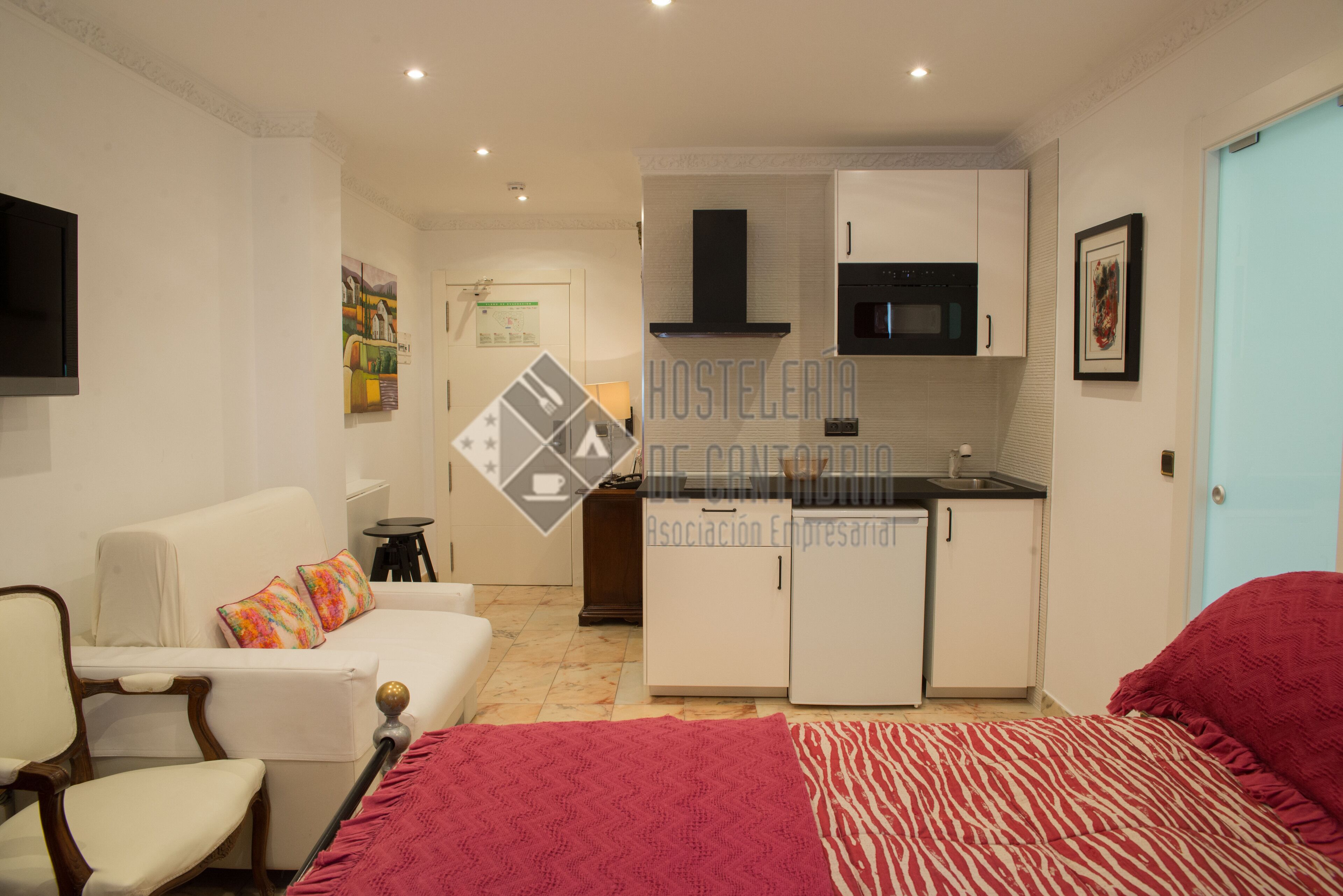 Habitación de hotel compacta con kitchenette, cama con colcha estampada en rojo y blanco, sofá blanco con almohadones coloridos y una pequeña silla de madera.