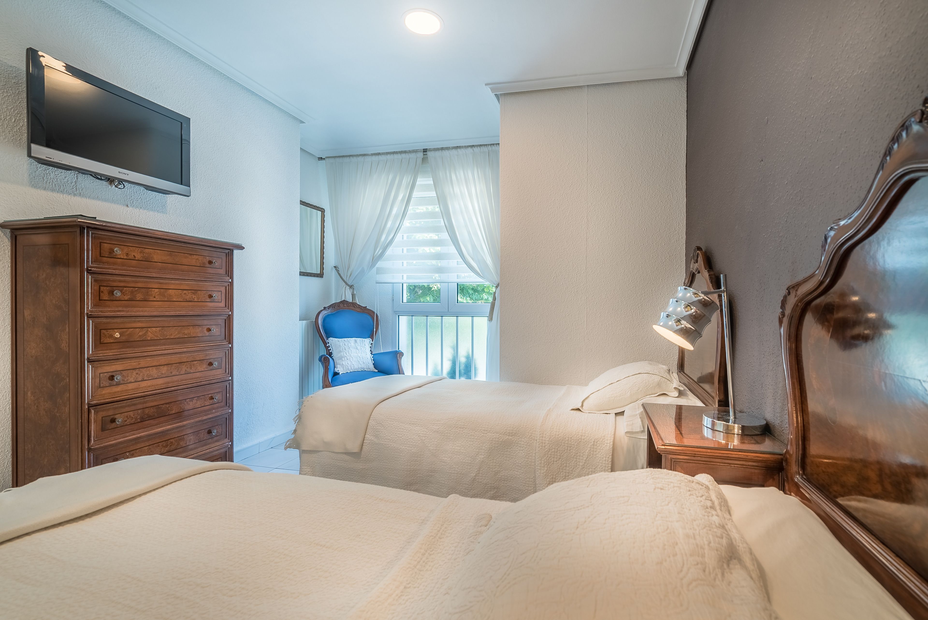 Un dormitorio con dos camas, un aparador, una silla, un televisor y una ventana con cortinas.