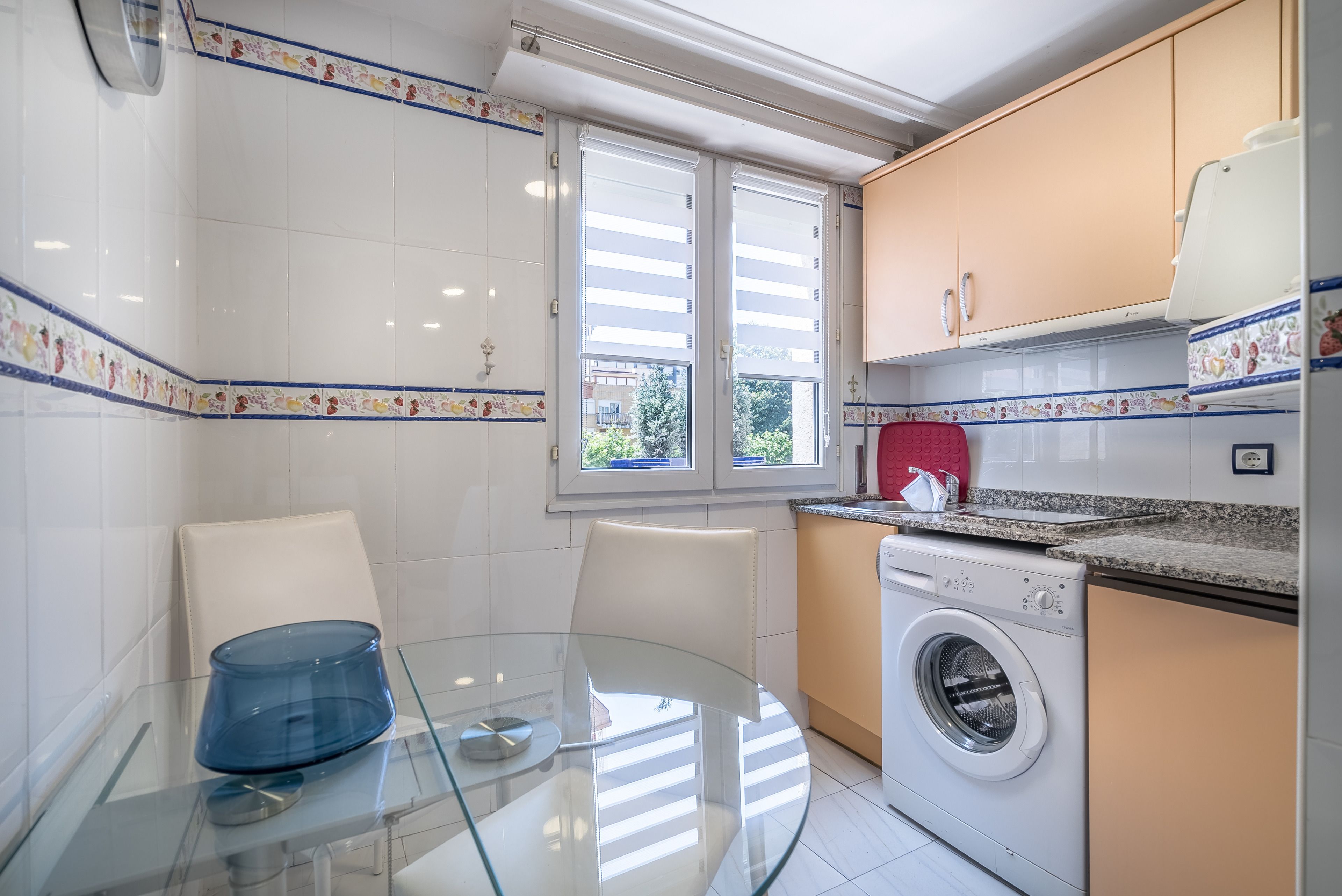 Een compacte keuken met een wasmachine, een glazen tafel en een raam met zonwering.
