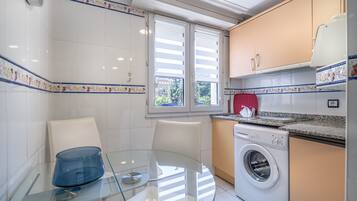 Een compacte keuken met een wasmachine, een glazen tafel en een raam met zonwering.