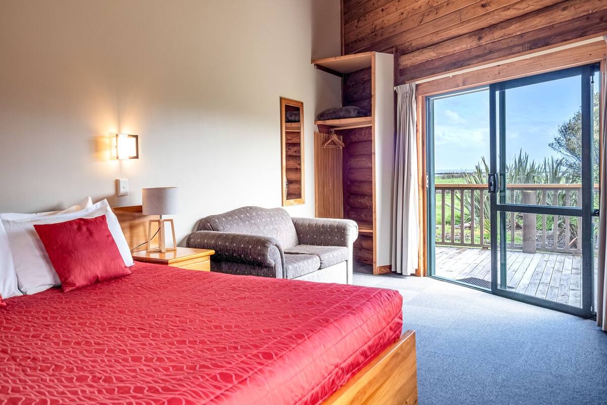 A bedroom with a large bed, a chair, a wooden wardrobe, and a view of the outdoors.