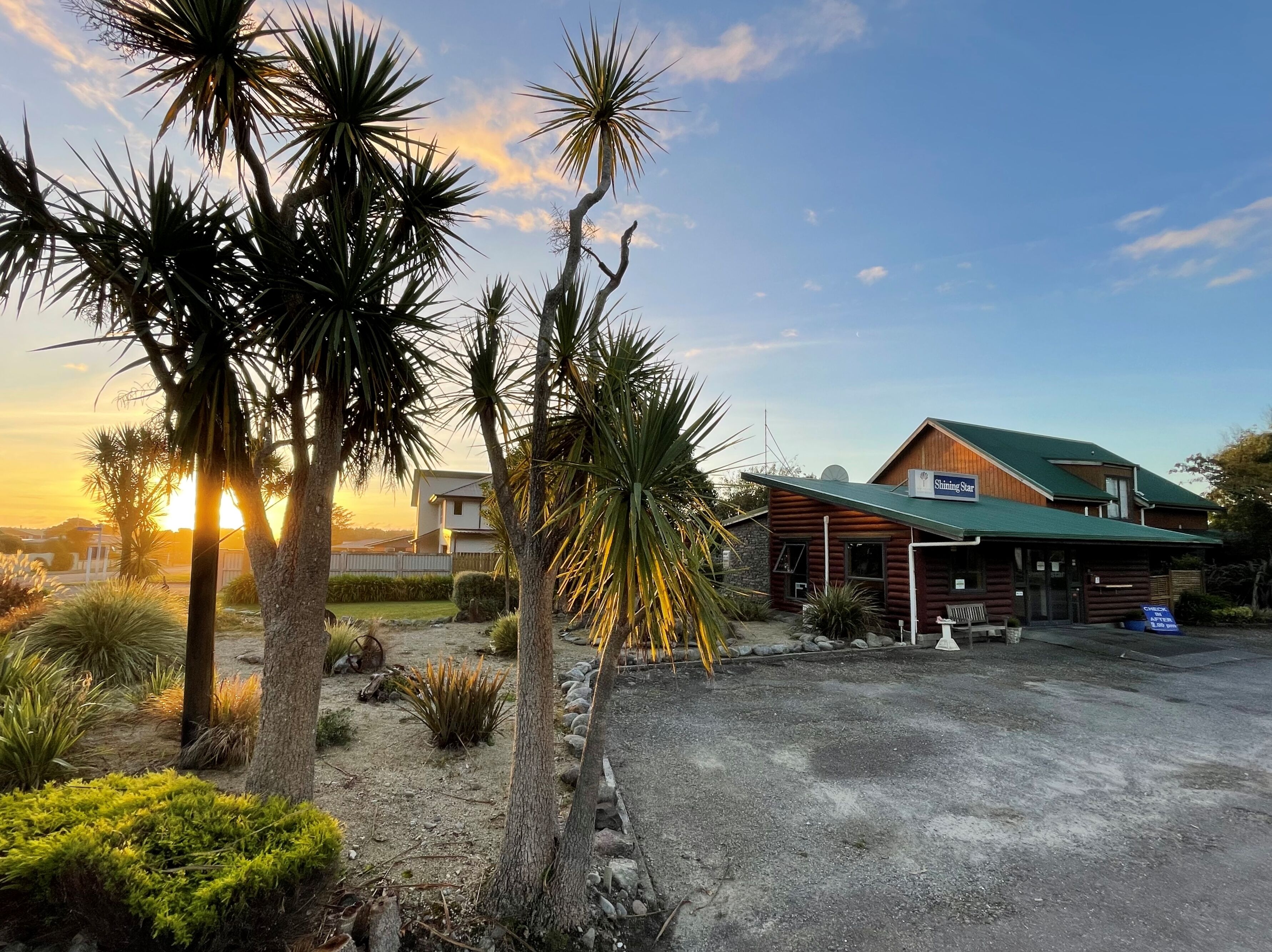 A rustic lodge with a green roof, surrounded by palm trees and a gravel driveway.