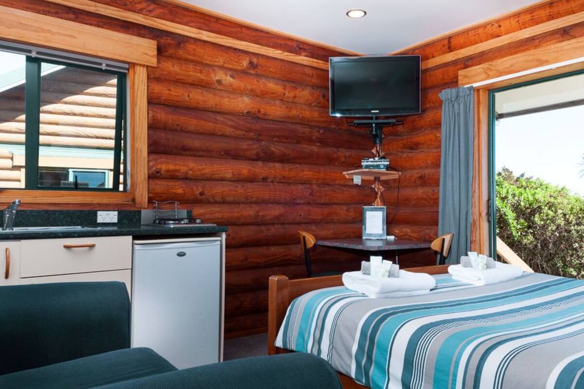 A cozy cabin room with a bed, a small desk, a television, and a window overlooking greenery.