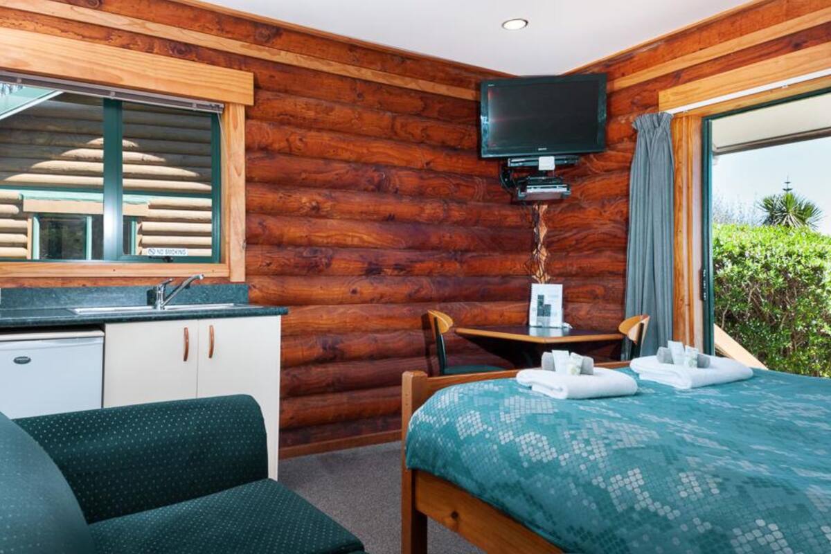 A room with a wooden log wall, a bed with a blue patterned bedspread, a small table with a lamp, a TV mounted on the wall, and a window with curtains.