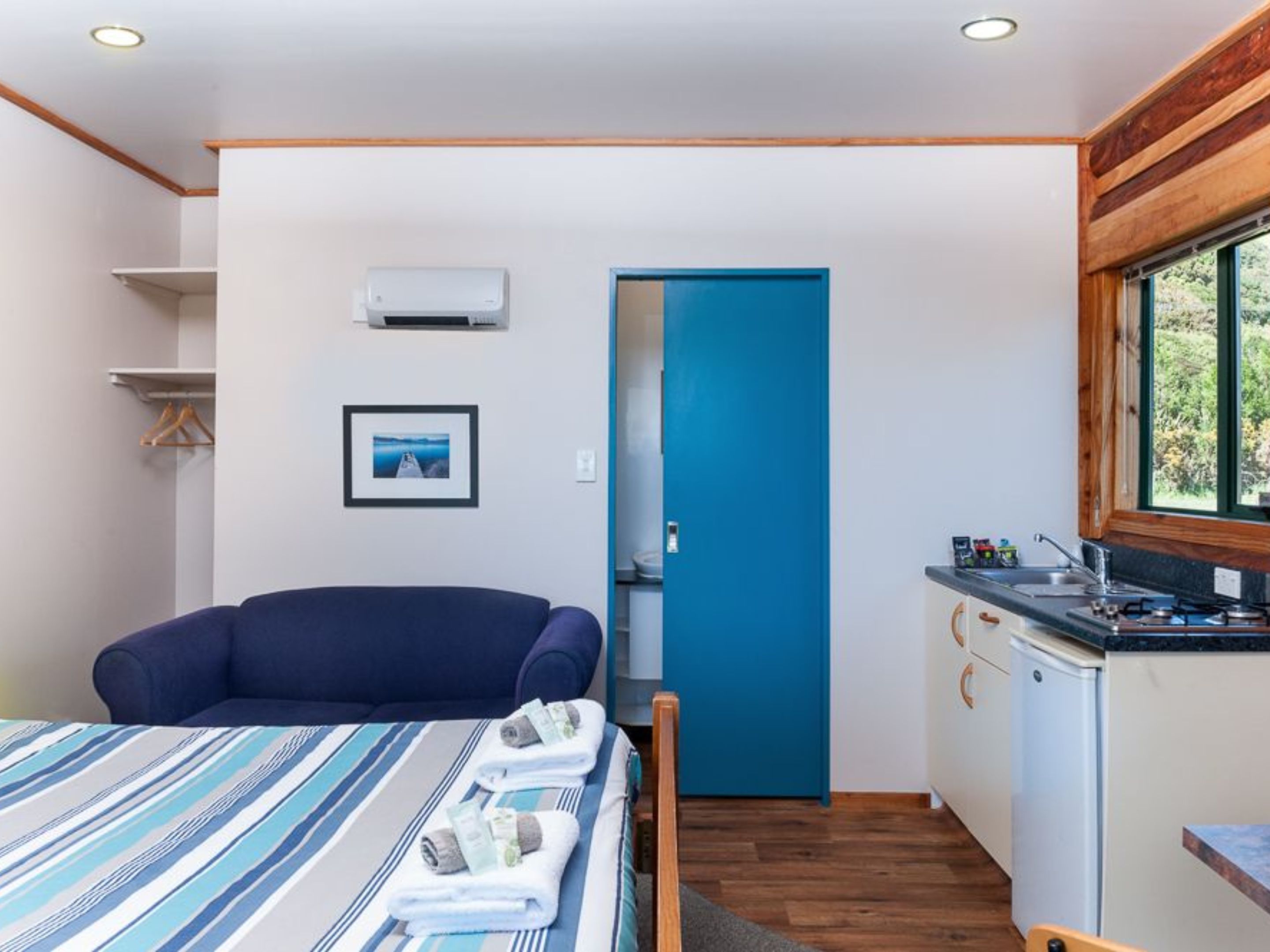 Una habitación pequeña con una cama, una puerta azul, una kitchenette y un cuadro en la pared.