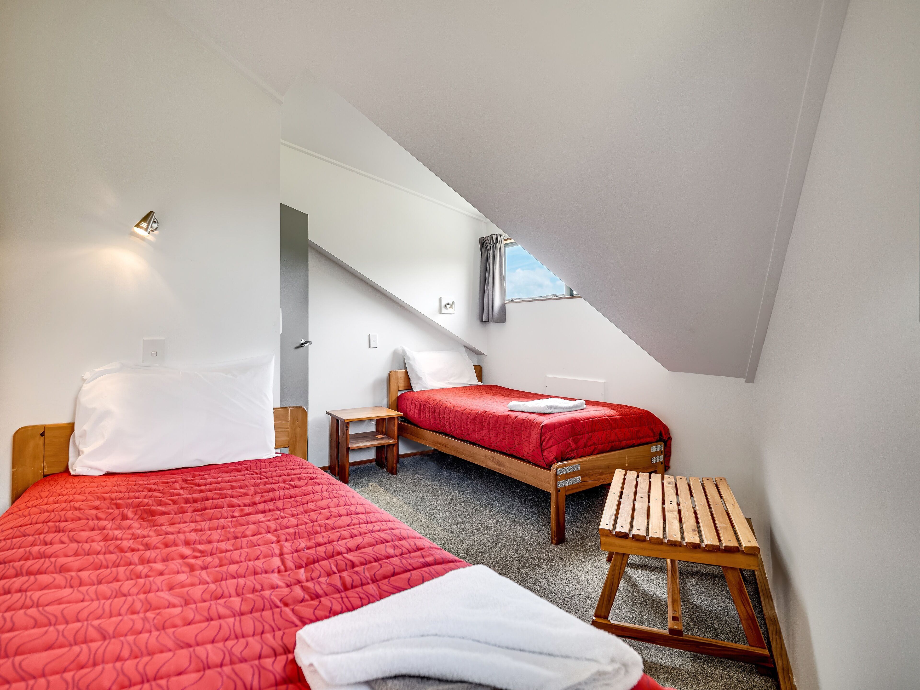A small, modern bedroom with two single beds, a wooden bench, and a skylight.