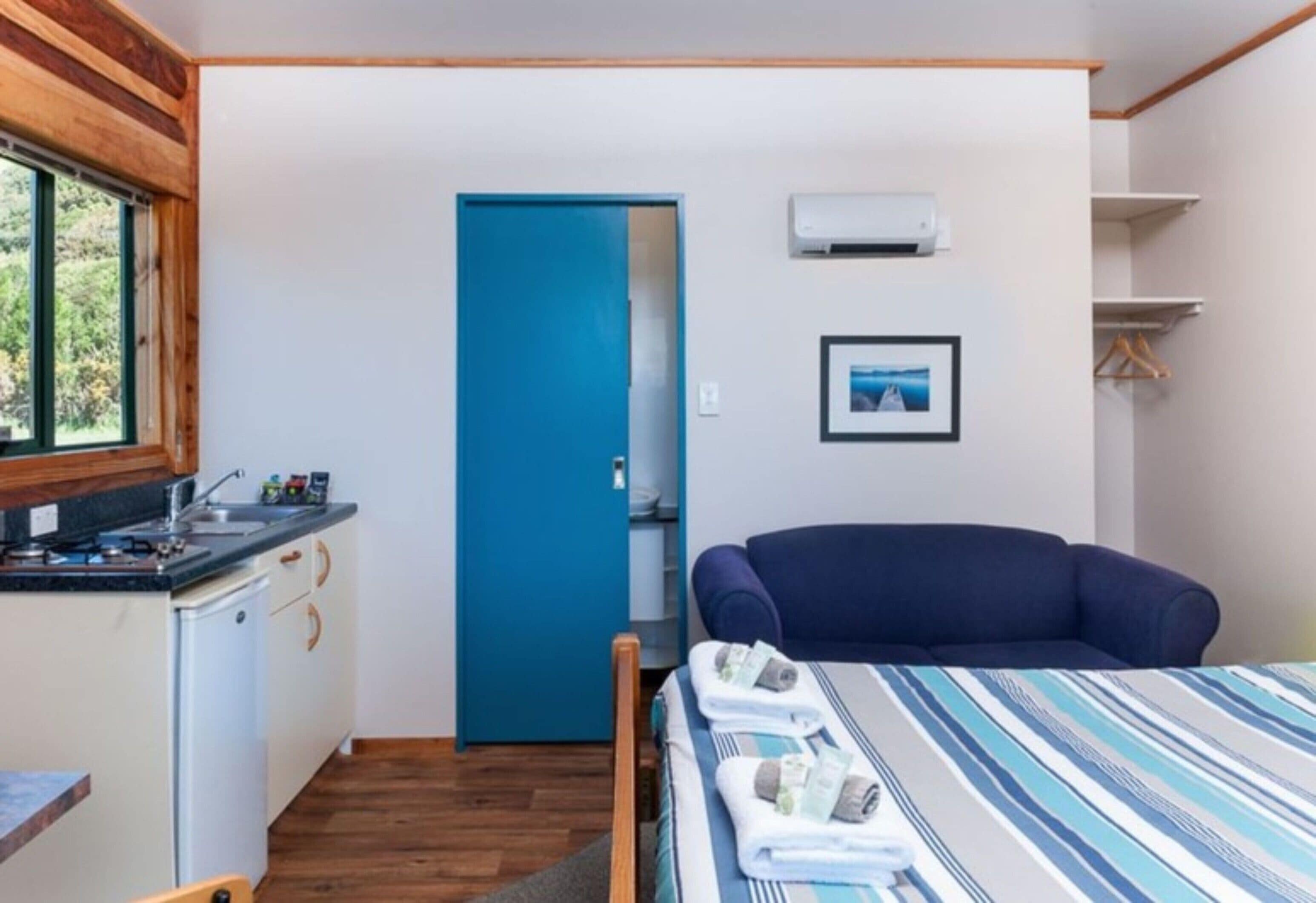 Quarto compacto com cozinha compacta, cama com roupa de cama listrada, porta azul e quadro na parede.