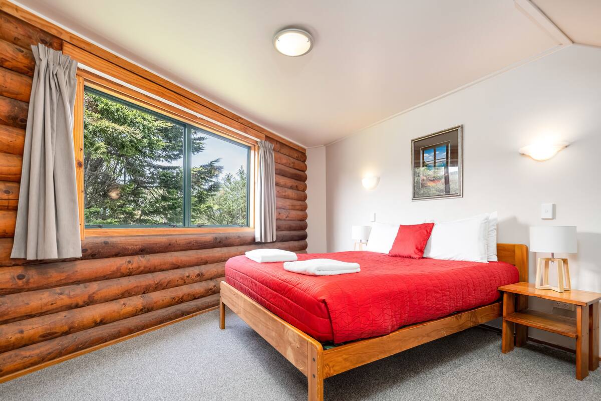 A log cabin bedroom with a wooden bed, a nightstand, a lamp, and a window with curtains.