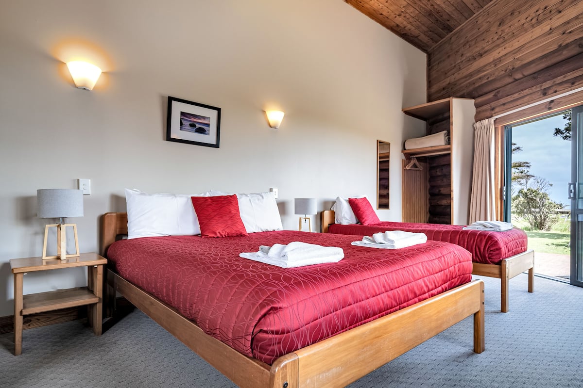A double bed with red bedding, two bedside tables with lamps, a framed picture on the wall, and a view through an open door.