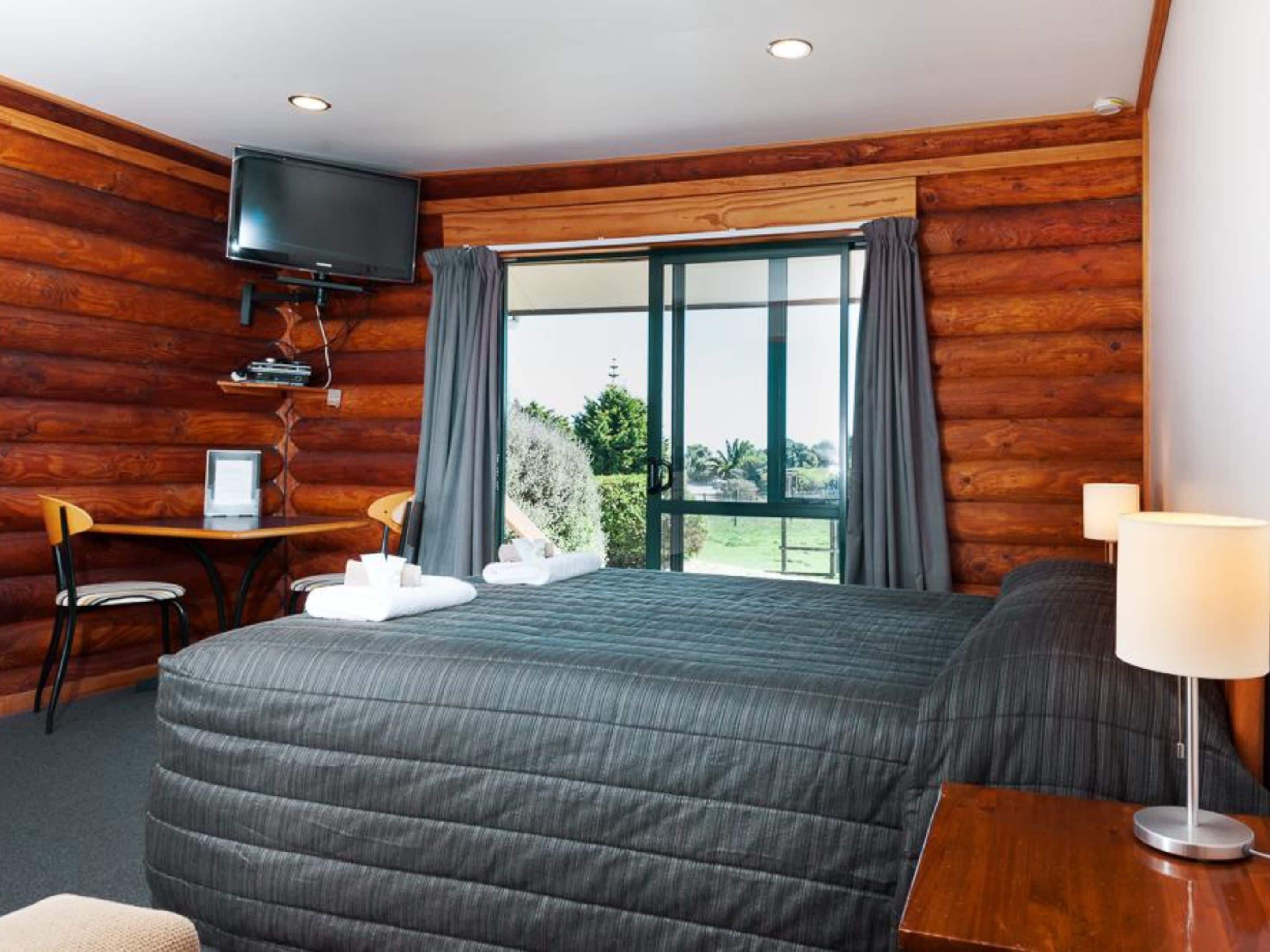 A log cabin-style room with a bed, a desk with a chair, a television, and a window with curtains.