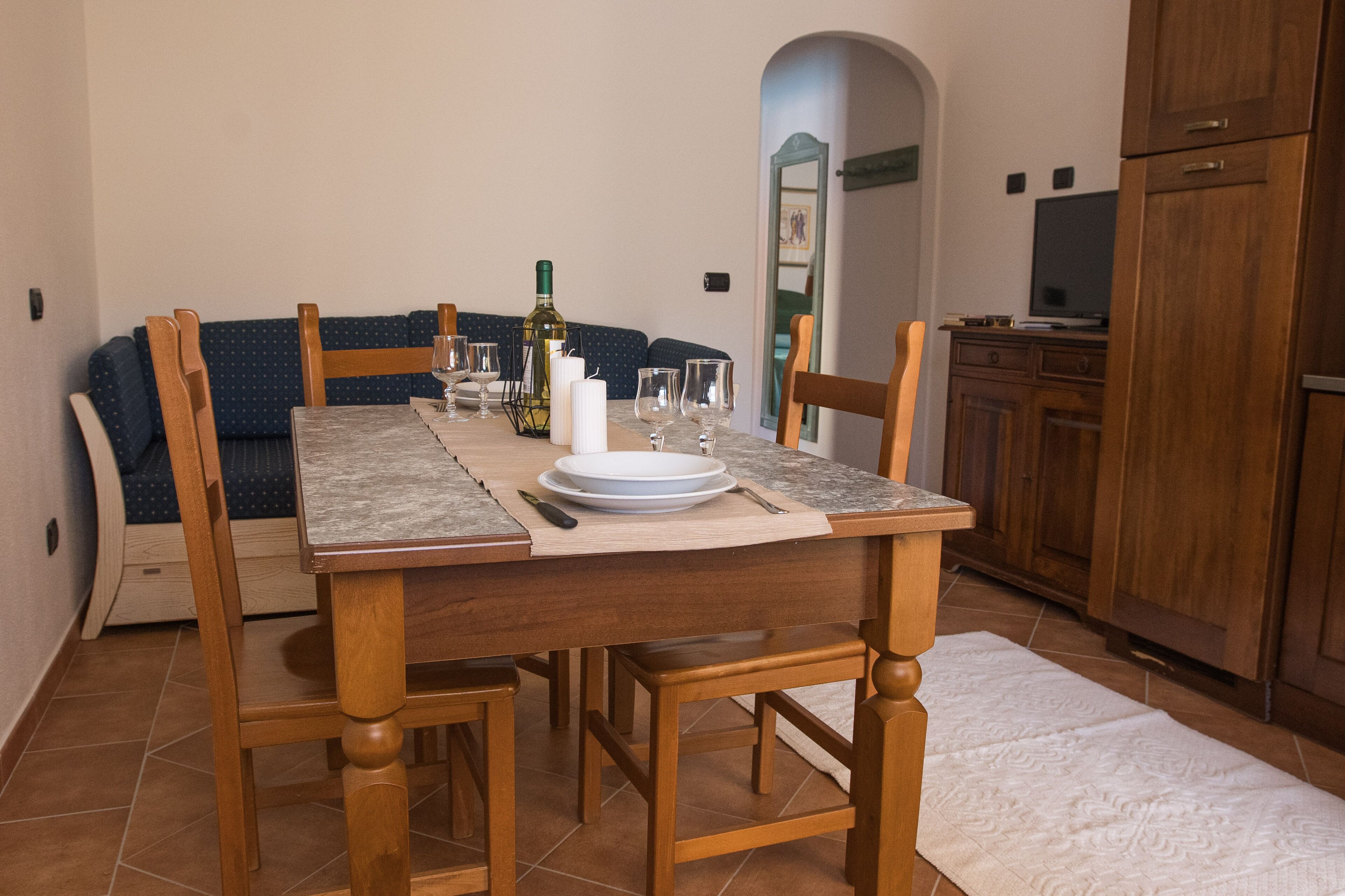Una mesa de comedor dispuesta con platos, vasos y una botella, rodeada de sillas de madera en una habitación con suelo de baldosas y un mueble de madera.