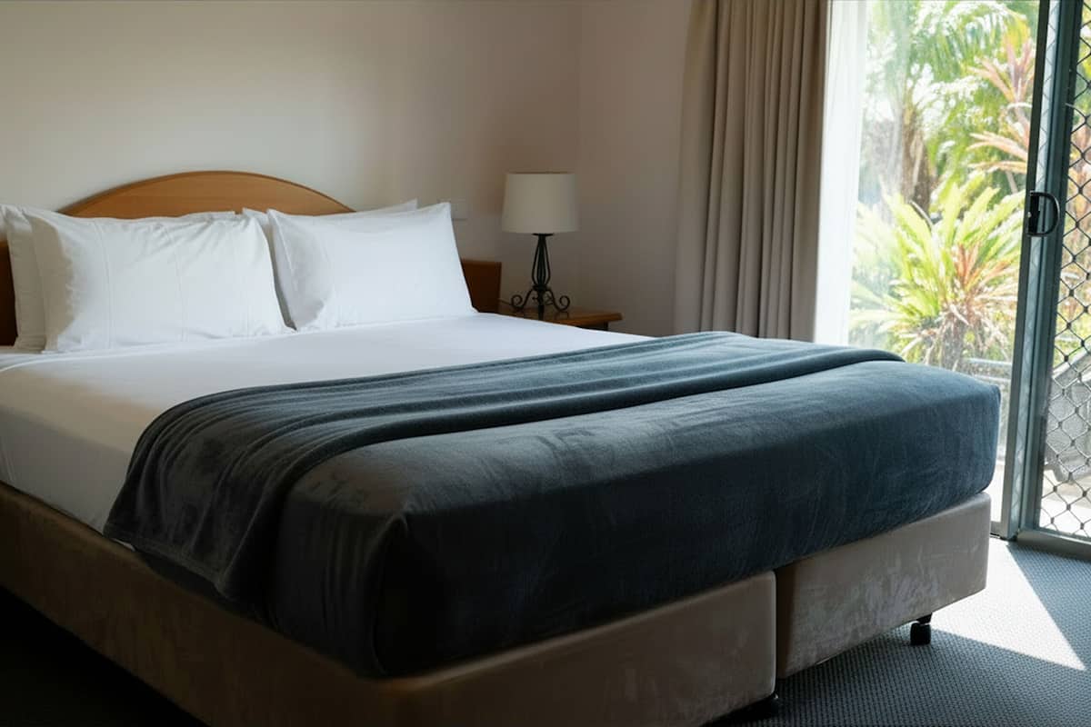 A hotel room with a bed, a nightstand, a lamp, and a view of greenery through the window.