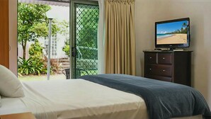 A hotel room with a bed, a TV on a dresser, and a sliding glass door leading to a balcony with a view of a beach.