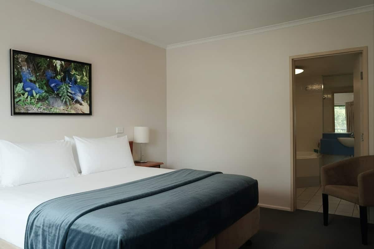 A hotel room with a bed, a desk, a chair, a painting, and a bathroom visible through an open door.
