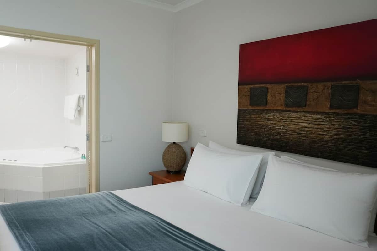 A hotel room with a bed, a nightstand with a lamp, a painting on the wall, and a bathroom visible through an open door.