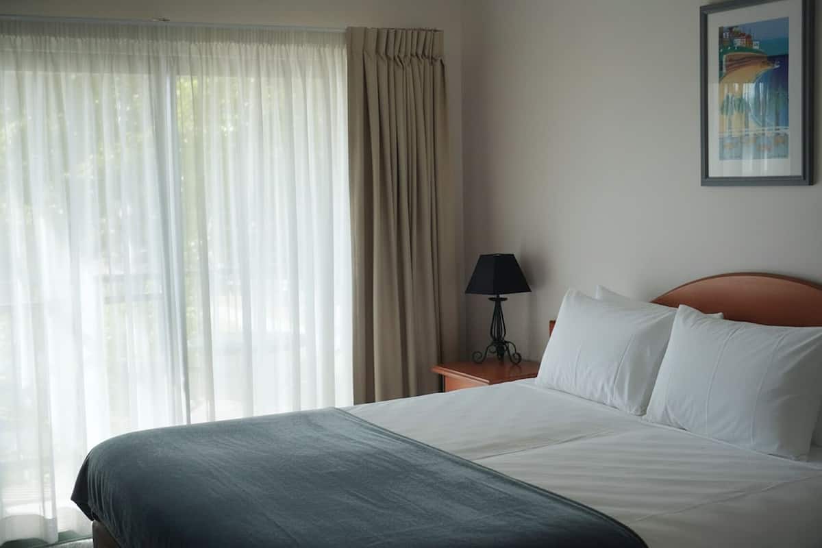 A hotel room with a bed, a nightstand with a lamp, a window with sheer curtains, and a framed picture on the wall.