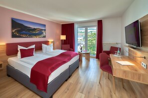 A hotel room with a large bed, a desk, a TV, and a balcony with a view of a lake and mountains. - Park Hotel (Traben-Trarbach)