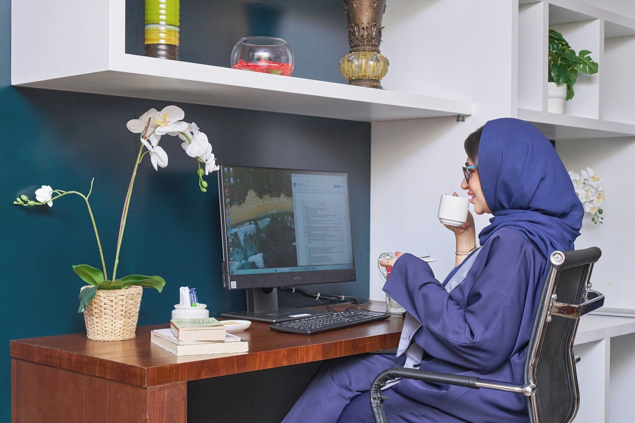 Una persona sentada en un escritorio con una computadora, un teclado, un ratón y una taza. Hay plantas y objetos decorativos en estanterías sobre el escritorio.