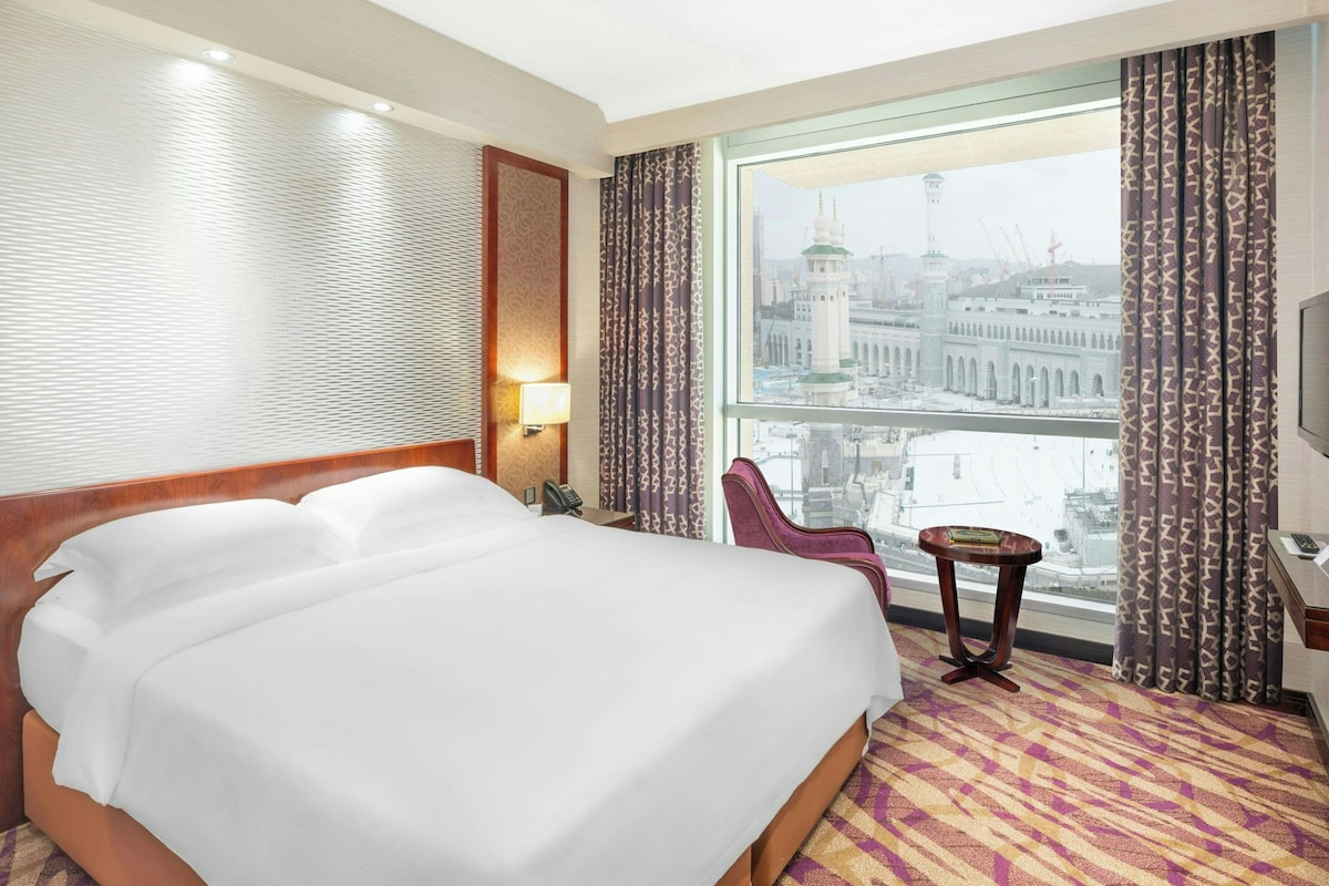 Une chambre d’hôtel avec un grand lit, une chaise, une petite table, une lampe et une vue sur le paysage urbain par la fenêtre.