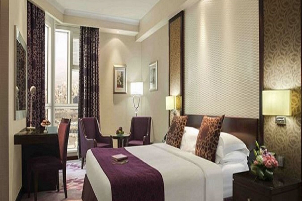 A hotel room with a large bed, a desk, two chairs, a small table, and a nightstand with a lamp.