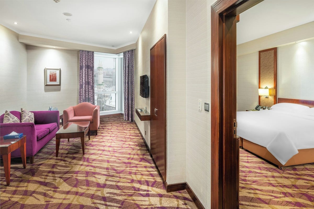 Una habitación de hotel con cama, sofá, sillón, televisor y ventana con cortinas.