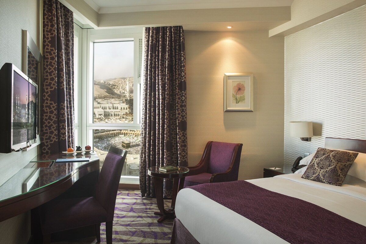 A hotel room with a bed, a desk, a chair, a small table, a TV, and a window with curtains.