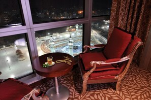 A room with a view of a large mosque, a red upholstered chair, a small table with a glass and a golden necklace, and patterned carpet. - Al Marwa Rayhaan by Rotana (Makkah)