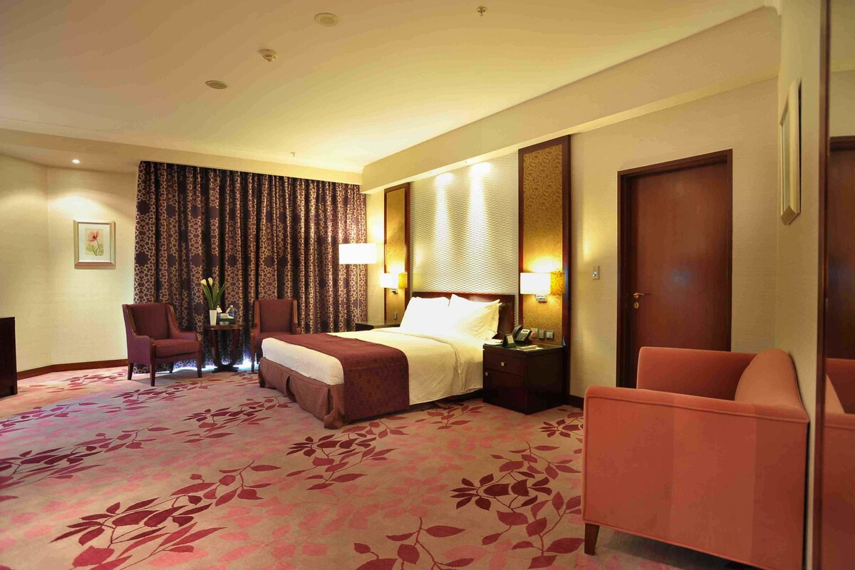 A hotel room with a large bed, two bedside tables with lamps, a chair, a desk with a vase, and a patterned carpet.