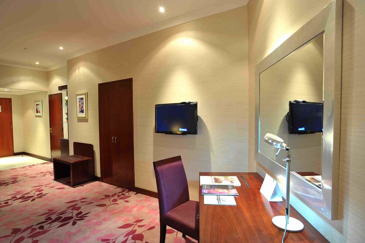 A hotel room with a large mirror, a desk with a lamp, a chair, a television, and a carpeted floor.