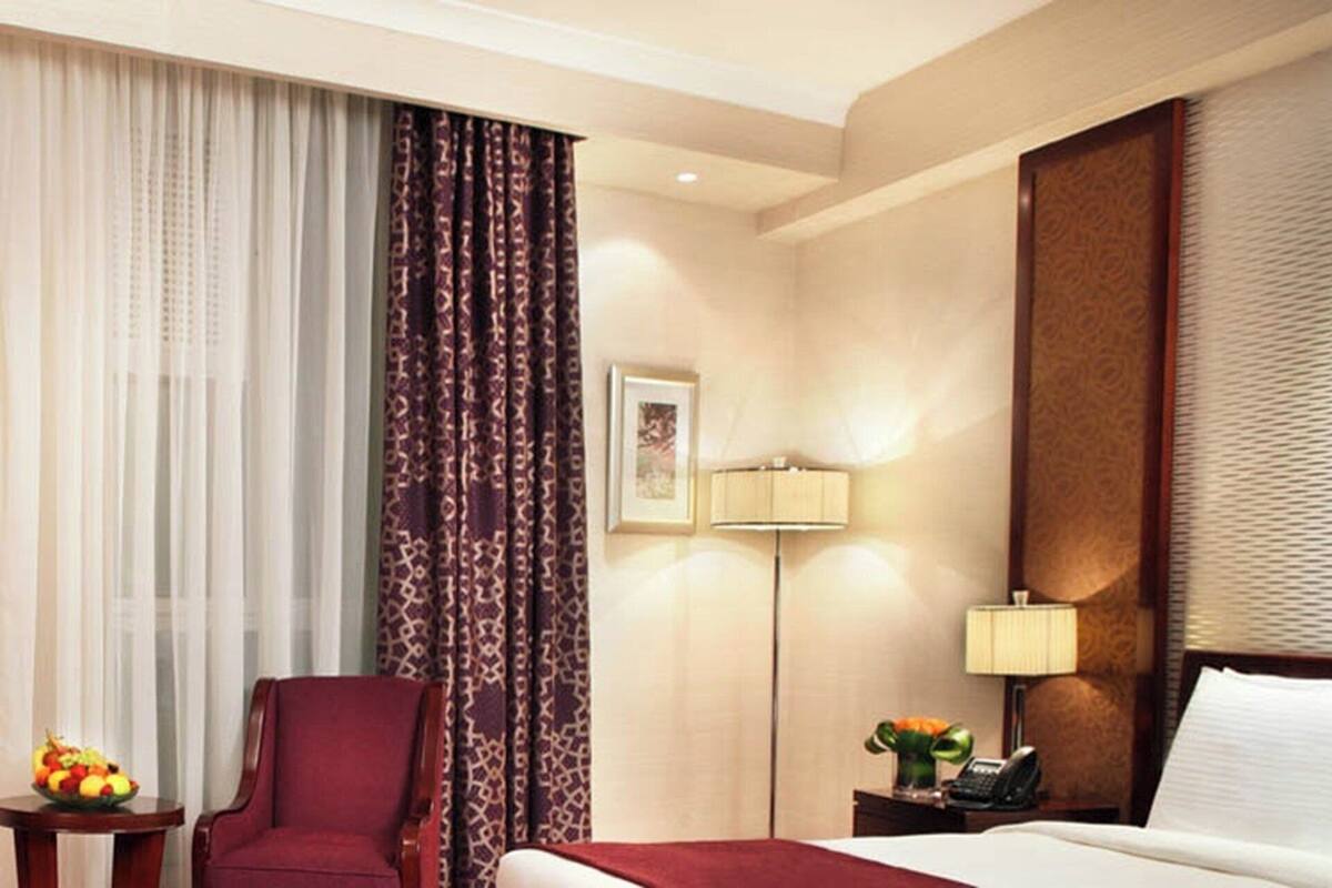 Habitación de hotel con una cama, un sillón rojo, una mesita con fruta, una lámpara y un teléfono.