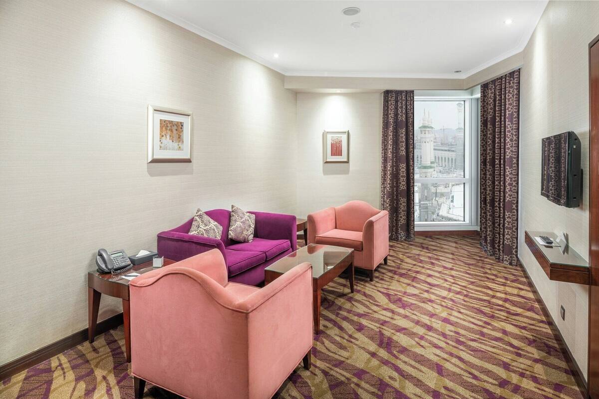 Habitación de hotel con un sofá púrpura, un sillón rosa, una pequeña mesa, un teléfono sobre una mesita auxiliar y un televisor montado en la pared.