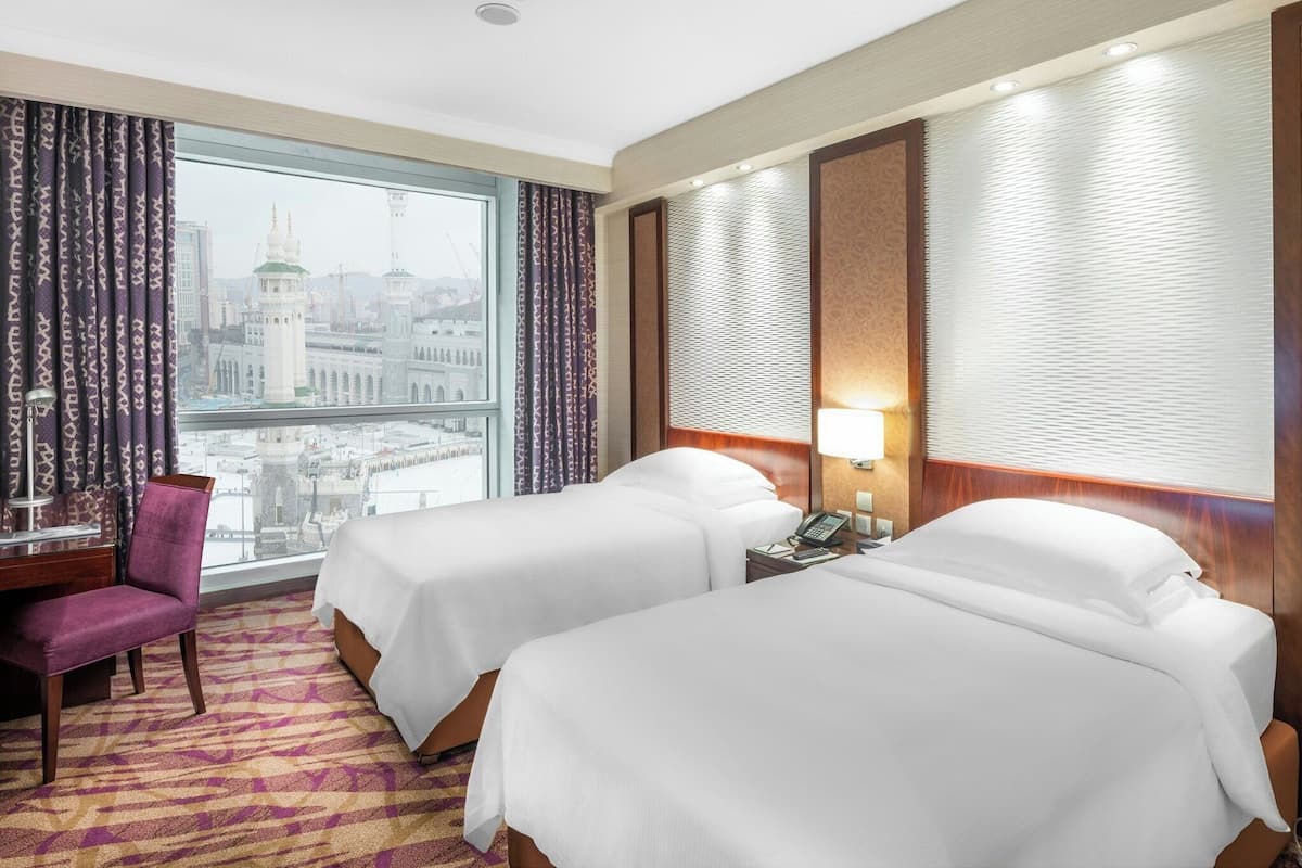 Habitación de hotel con dos camas, un escritorio, una silla y un amplio ventanal con vistas a un paisaje urbano.