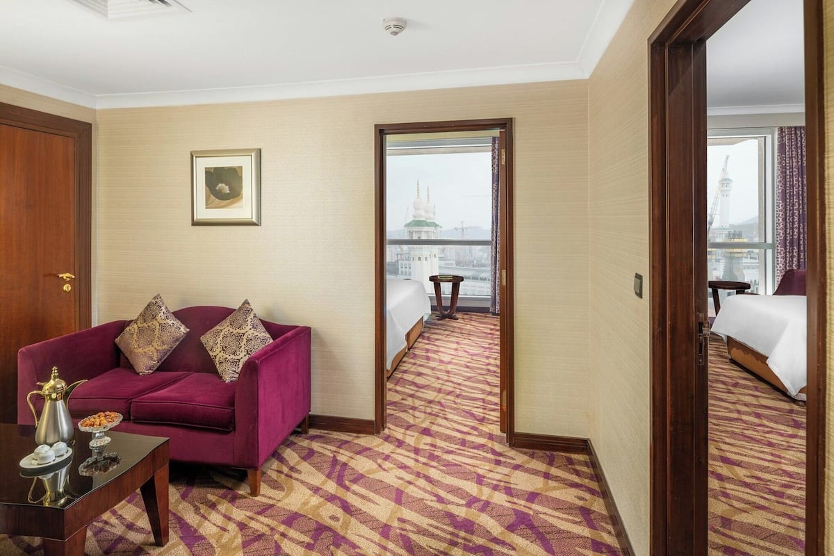 Una habitación de hotel con un sofá morado, una mesita con una tetera y una cama con una colcha blanca. Un cuadro enmarcado cuelga de la pared, y una ventana ofrece vistas a un edificio con una cúpula verde.