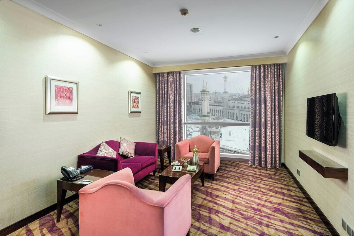 Habitación de hotel con un ventanal grande, un sofá, una silla, una mesa de centro y un televisor de pantalla plana.