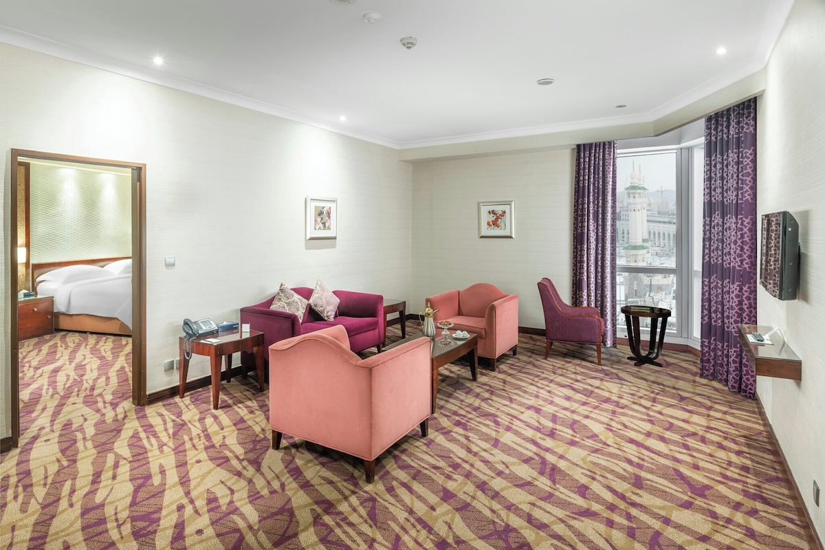 Habitación de hotel con un ventanal grande, una cama, dos sillones rosados, una mesita y un televisor en la pared.