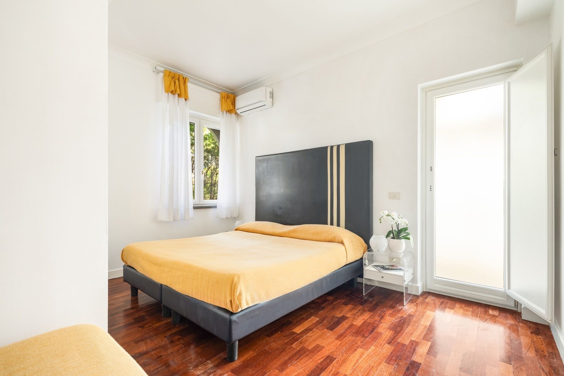 Un dormitorio con una cama, una mesita de noche, una planta en maceta y un ventanal con cortinas.