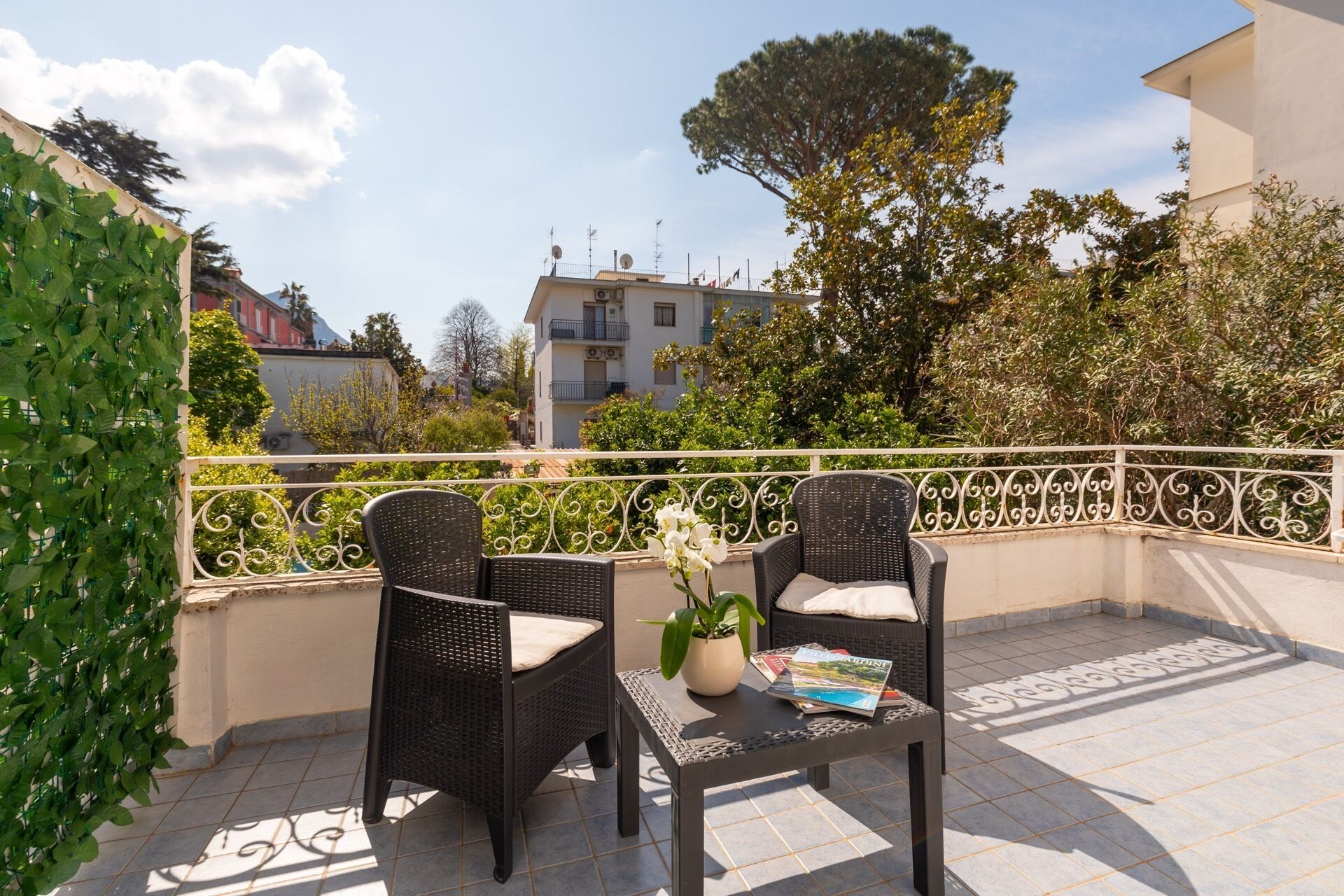 A balcony with two wicker chairs and a small table, surrounded by a white railing and greenery.