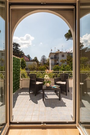 Ein Balkon mit zwei Korbstühlen, einem Tisch, einer Topfpflanze und Blick auf Häuser und Bäume.